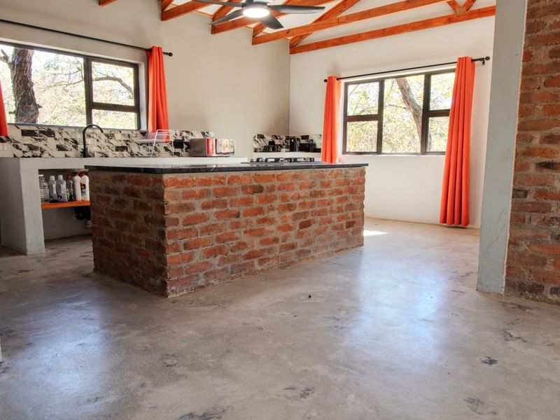 Open-plan kitchen featuring exposed brickwork, timber roof trusses, concrete flooring, and a central brick island, offering a rustic industrial aesthetic.
