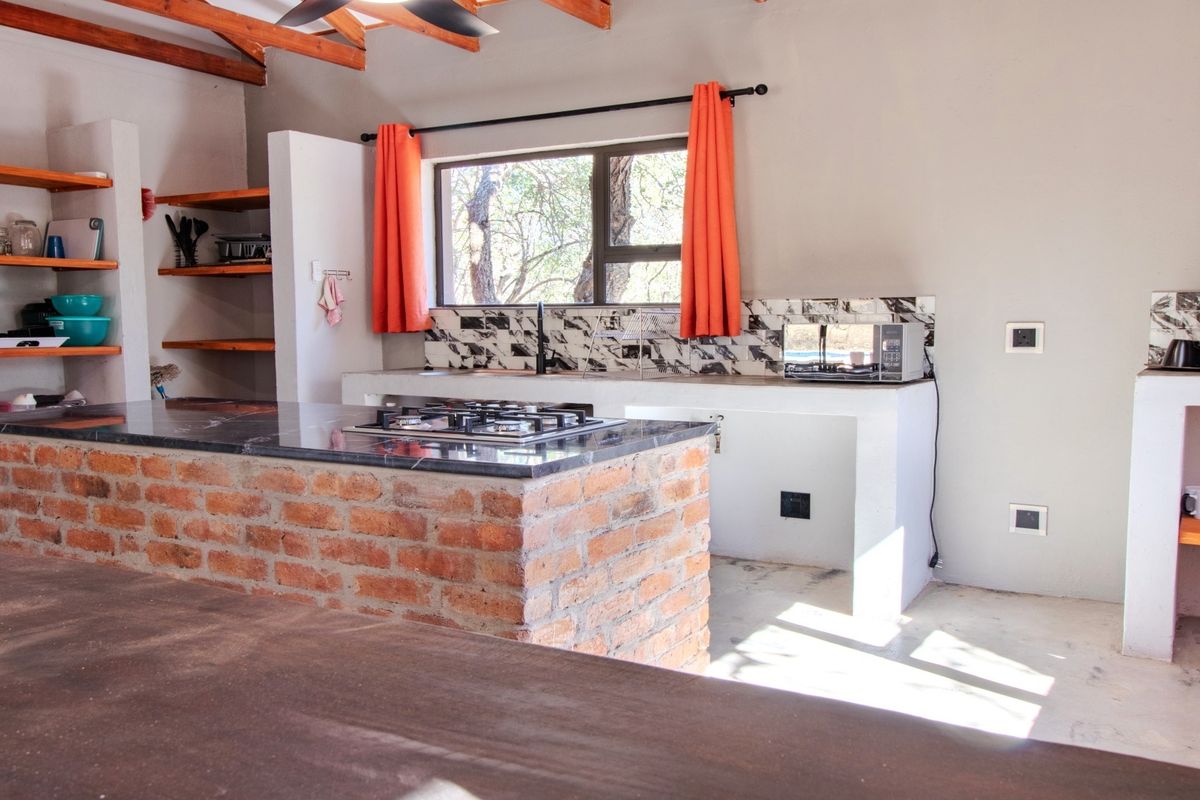 Rustic kitchen featuring an exposed brick island, stone countertops, built-in shelving, and exposed timber ceiling beams with a tiled backsplash.