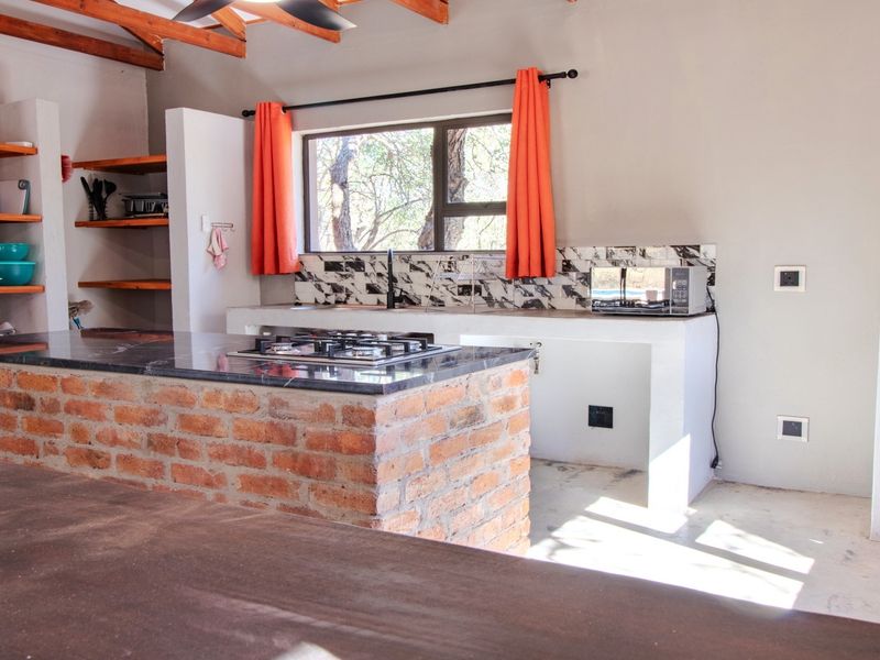 Rustic kitchen featuring an exposed brick island, stone countertops, built-in shelving, and exposed timber ceiling beams with a tiled backsplash.