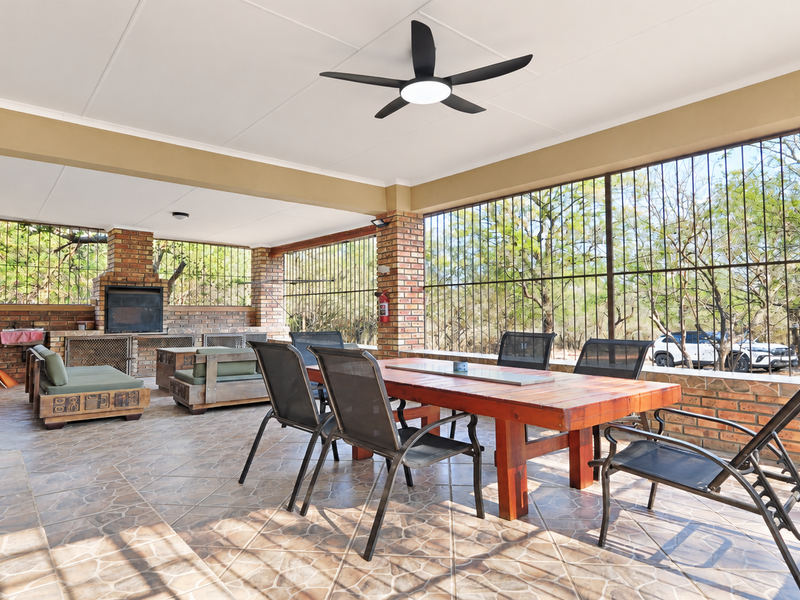 Spacious covered patio featuring a built-in braai, tiled flooring, and ceiling fan, enclosed with security bars overlooking the garden.