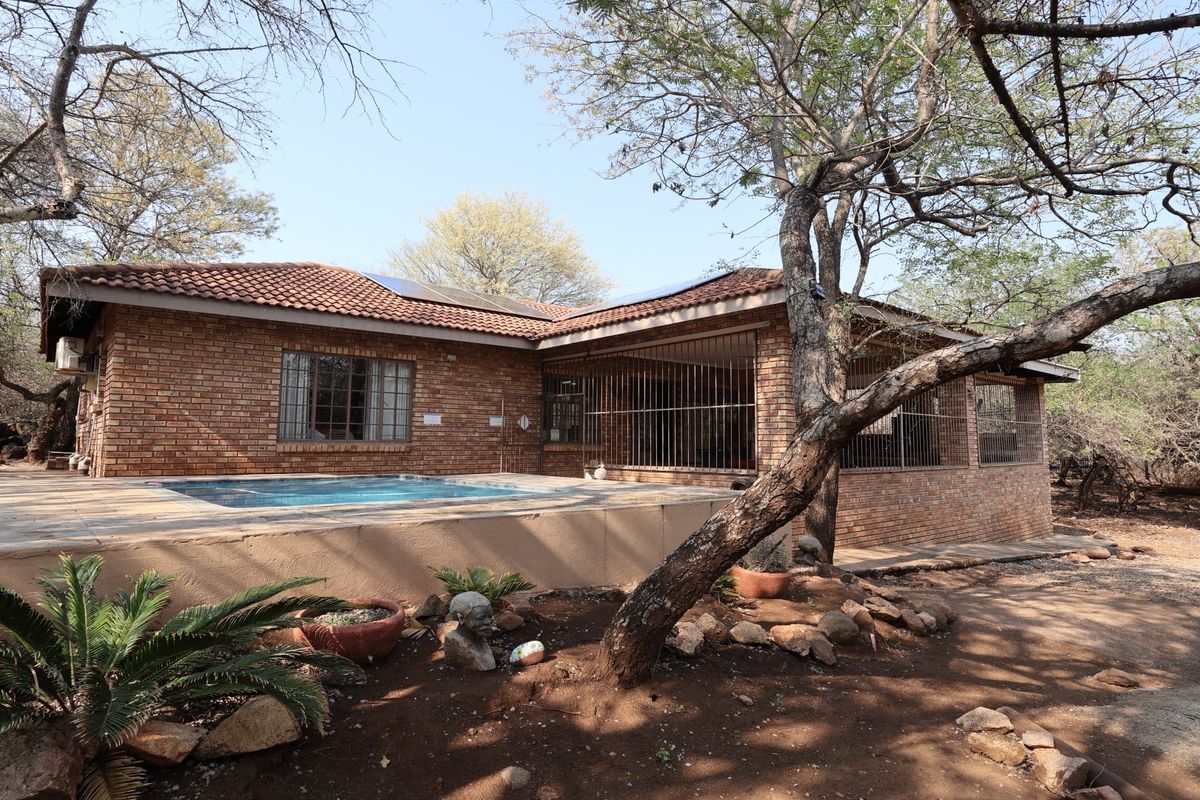 Rear view of a brick house featuring a swimming pool, security bars on windows, and solar panels set within a natural bushveld environment.