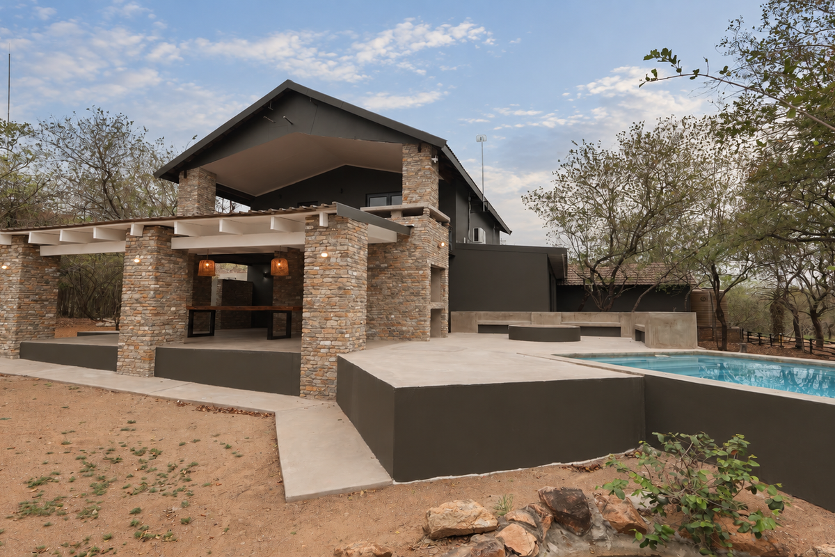 Rear exterior view featuring a covered patio with stone pillars, built-in braai, concrete seating area, and a swimming pool set in a natural landscape.