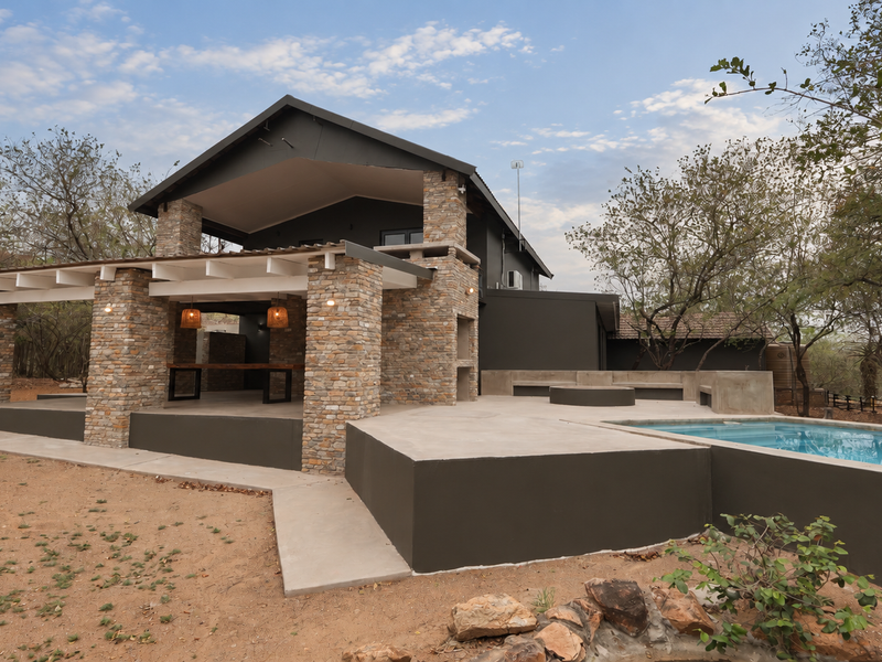 Rear exterior view featuring a covered patio with stone pillars, built-in braai, concrete seating area, and a swimming pool set in a natural landscape.