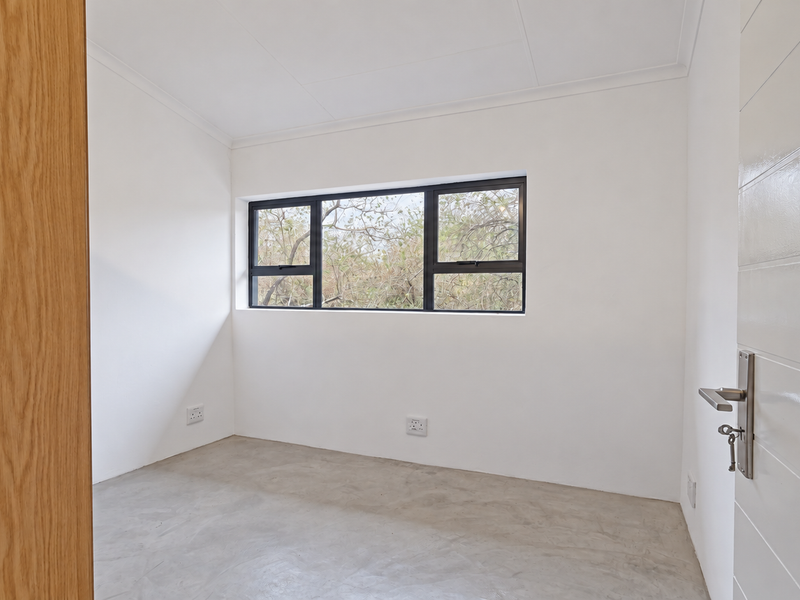 A bright, empty bedroom featuring polished concrete floors, white walls, and black aluminium window frames with a view of the surrounding trees.