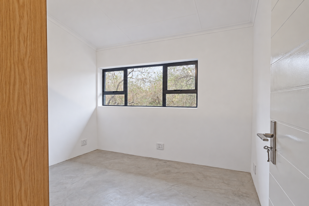 A bright, empty bedroom featuring polished concrete floors, white walls, and black aluminium window frames with a view of the surrounding trees.