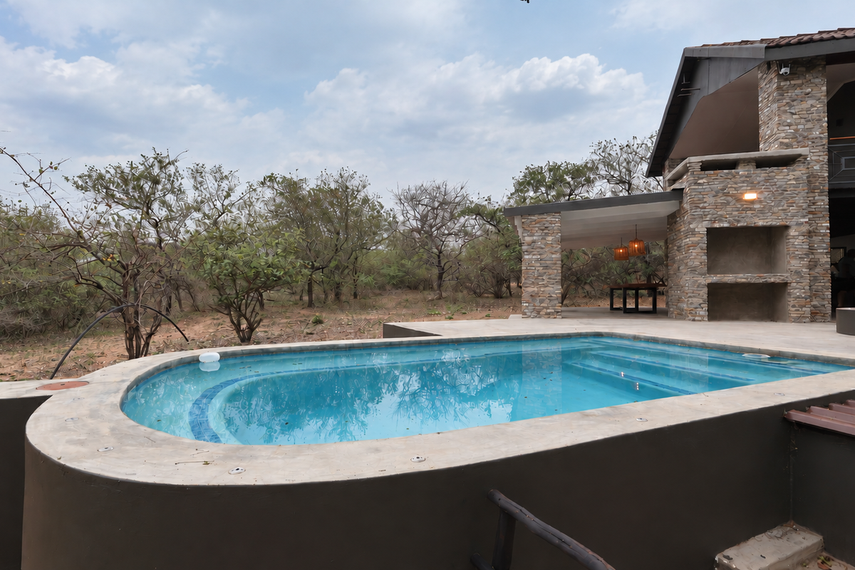 Outdoor pool area featuring a swimming pool, stone-clad exterior walls, and a covered patio with a built-in braai, set against a natural bushveld backdrop.