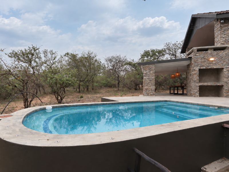 Outdoor pool area featuring a swimming pool, stone-clad exterior walls, and a covered patio with a built-in braai, set against a natural bushveld backdrop.
