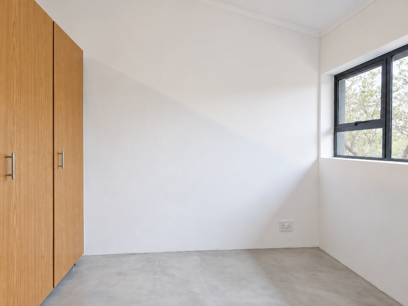 A bright, empty bedroom featuring modern concrete flooring, built-in wooden cupboards, and a large window with a view of the surrounding trees.