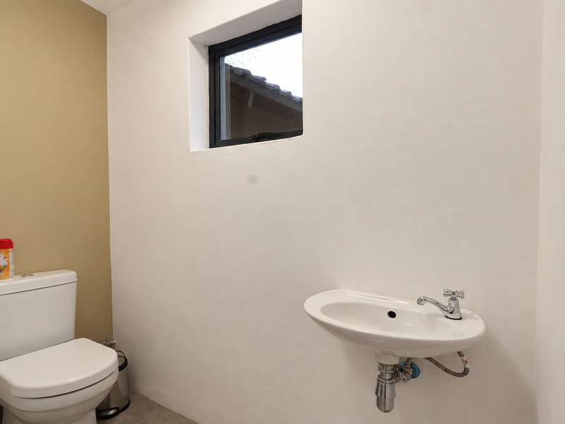 A clean, minimalist bathroom featuring a wall-mounted basin with chrome fixtures and a standard toilet, illuminated by a small casement window.