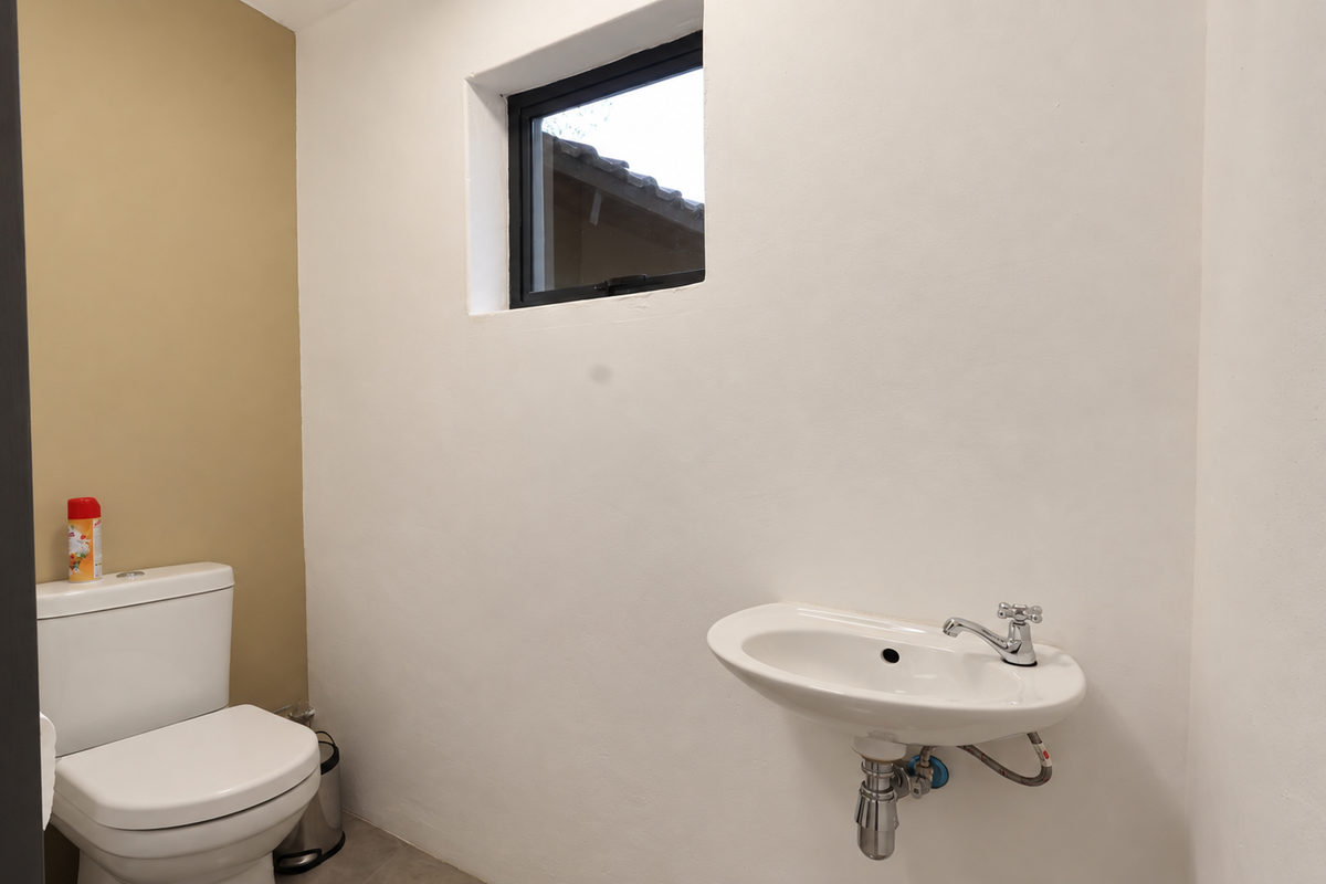 A clean, minimalist bathroom featuring a wall-mounted basin with chrome fixtures and a standard toilet, illuminated by a small casement window.
