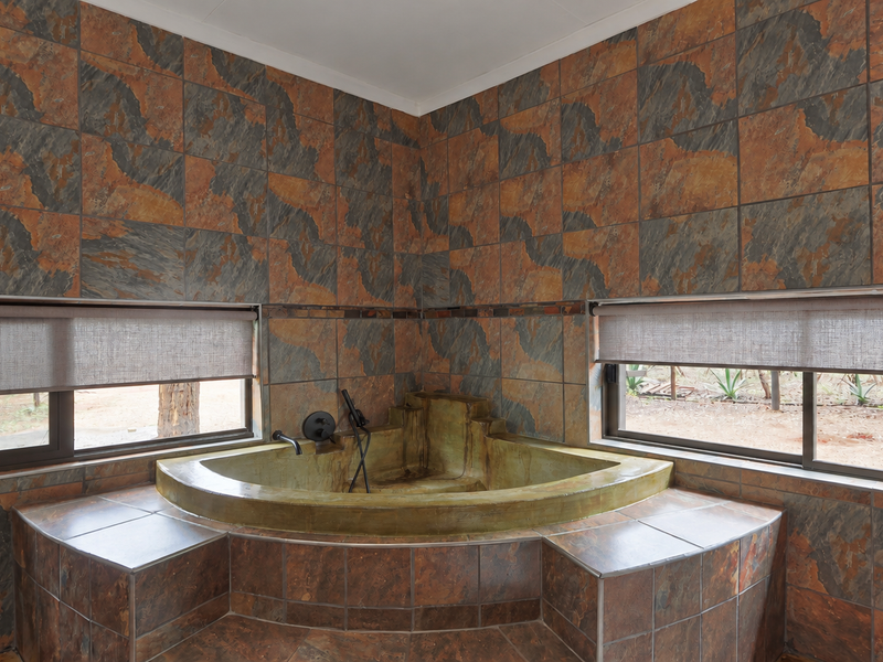 A bathroom featuring a built-in corner bathtub surrounded by rustic slate tiling and large windows with roller blinds.