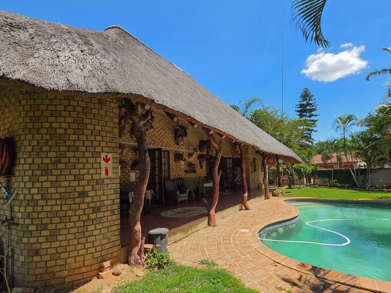 Rear exterior view featuring a thatched roof house, a brick-paved patio area, and a swimming pool set within a landscaped garden.