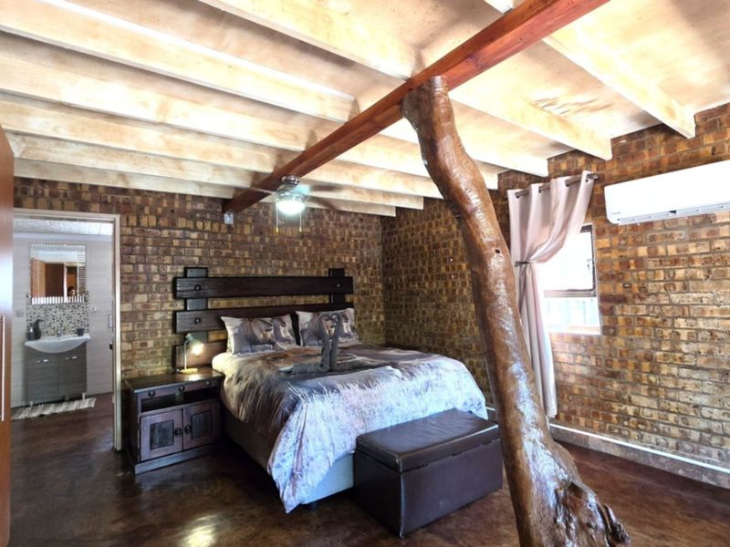 A rustic bedroom featuring exposed brick walls, timber ceiling beams, a split-system air conditioner, and an en-suite bathroom visible through the doorway.