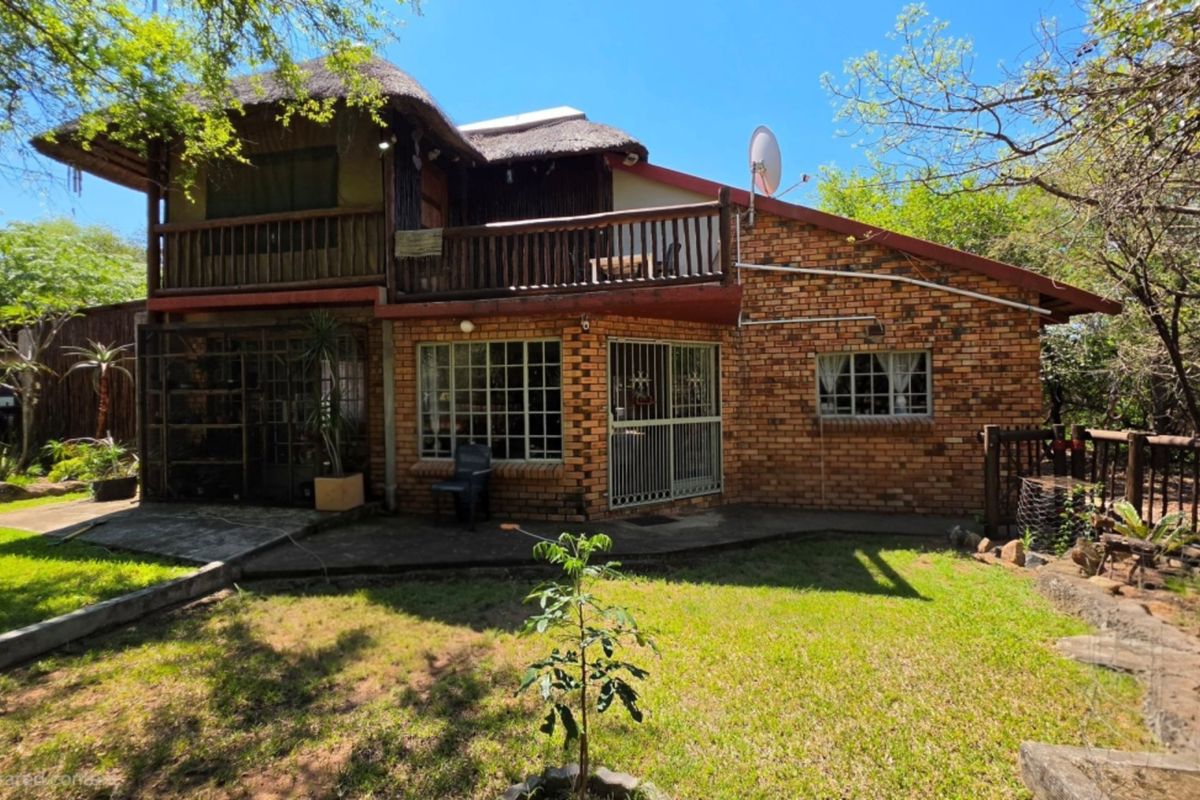 Rear view of a double-story brick house featuring a thatched roof, a wooden balcony, and a surrounding garden area.