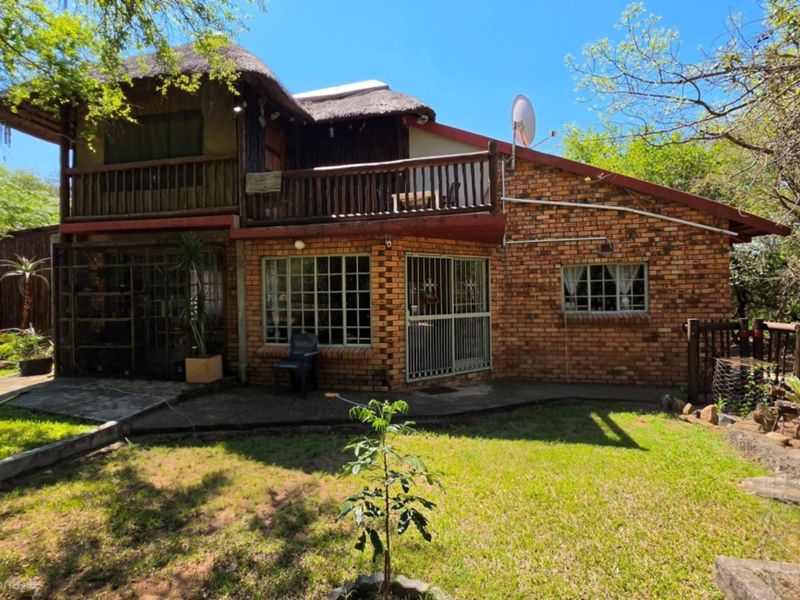Rear view of a double-story brick house featuring a thatched roof, a wooden balcony, and a surrounding garden area.