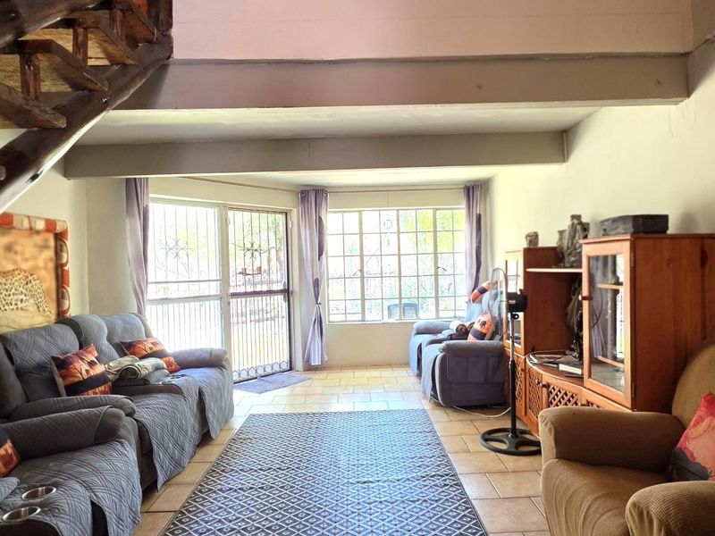 A living room featuring tiled flooring, exposed timber staircase, and natural light through steel-framed windows with security access.