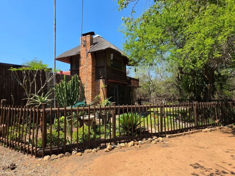 A rustic house exterior featuring a thatched roof, brick chimney, and timber balcony, surrounded by a fenced garden in a natural bushveld setting.