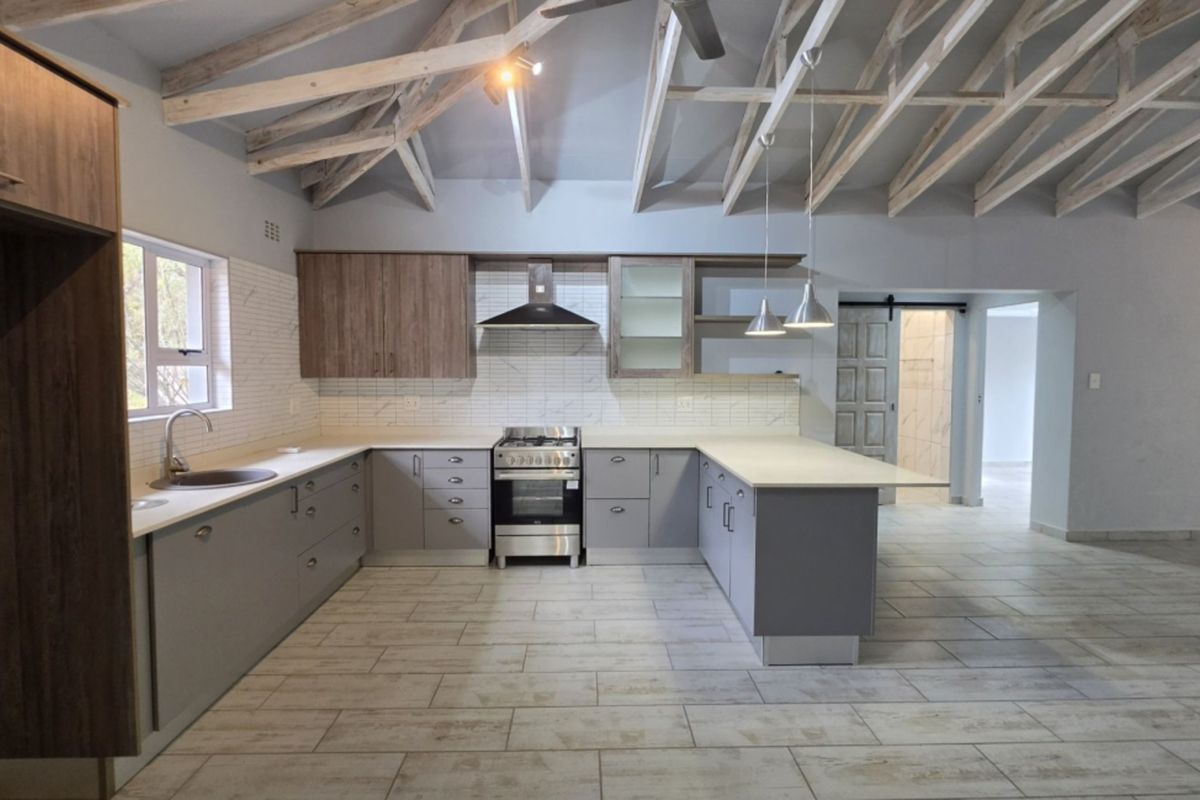 Modern open-plan kitchen featuring exposed timber trusses, sleek grey cabinetry, stone countertops, stainless steel appliances, and tiled flooring in a bright, spacious layout.