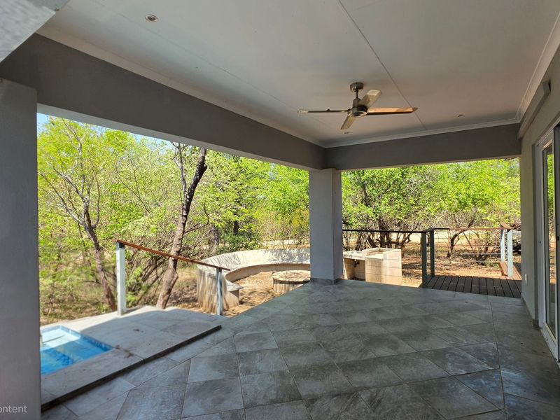 Spacious covered patio featuring tiled flooring, a ceiling fan, built-in braai, and integrated seating, overlooking a natural garden setting.