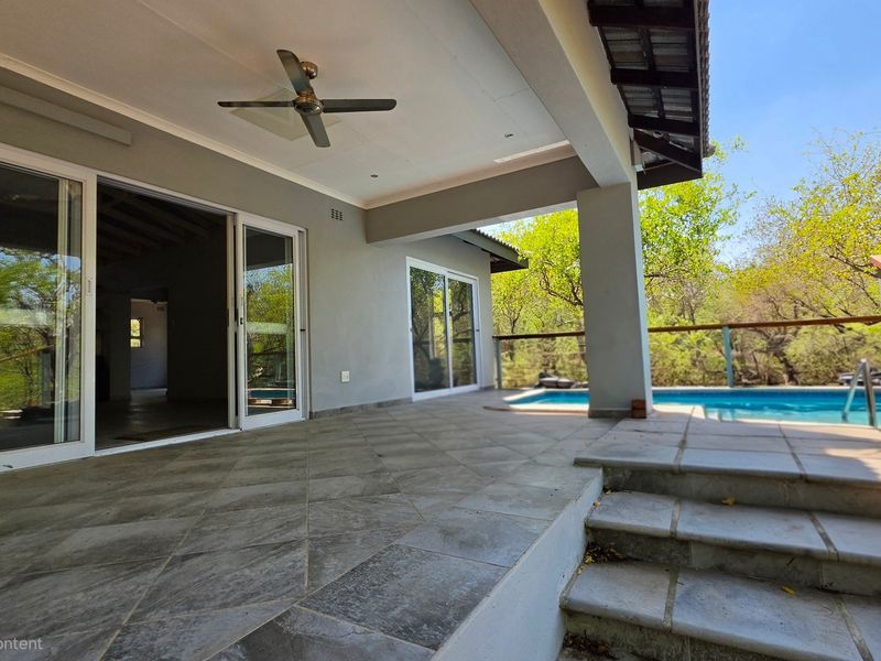 A spacious covered patio featuring tiled flooring and a ceiling fan, providing seamless access to the swimming pool and surrounding natural landscape.