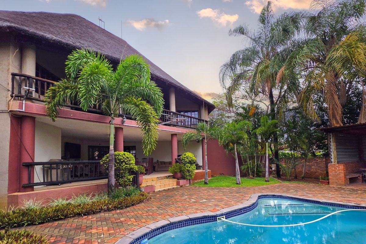 Rear exterior view featuring a thatched roof house, covered veranda, brick paved patio, and a swimming pool set within a landscaped garden.