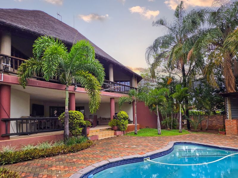 Rear exterior view featuring a thatched roof house, covered veranda, brick paved patio, and a swimming pool set within a landscaped garden.