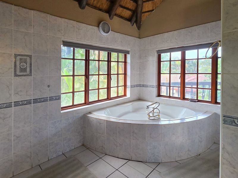 A spacious bathroom featuring a corner bathtub, tiled finishes, and large timber-framed windows under a traditional thatched roof.