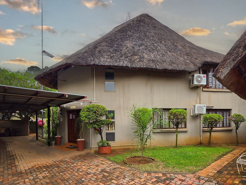 Rear view of a house featuring a traditional thatch roof, paved driveway, carport, and established garden area with exterior air conditioning units.