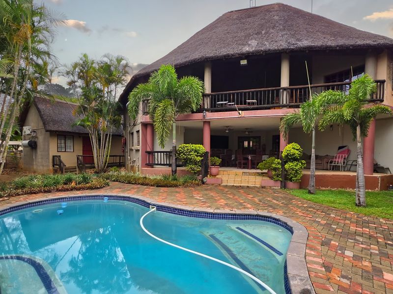 A spacious outdoor area featuring a swimming pool, a large thatched-roof house with a covered patio, and a balcony overlooking the garden.