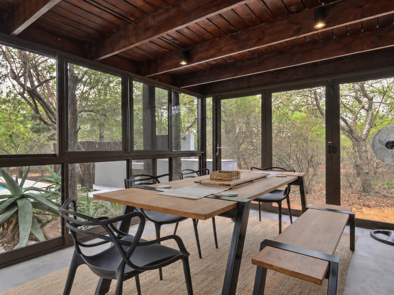 A covered patio area featuring exposed timber beam ceilings, large glass sliding doors, and concrete flooring, offering a seamless transition to the outdoor garden space.