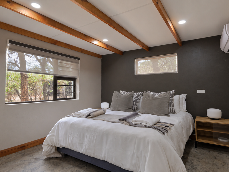A modern bedroom featuring exposed timber ceiling beams, polished concrete floors, built-in cabinetry, and a wall-mounted split-system air conditioner.