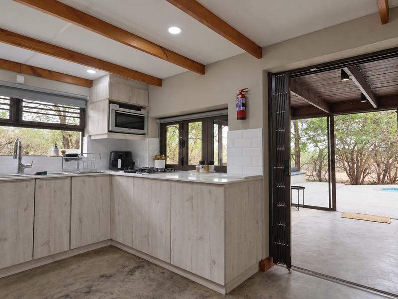 Modern kitchen featuring light wood cabinetry, stone countertops, subway tile backsplash, and exposed timber beams, with seamless access to an outdoor patio area.