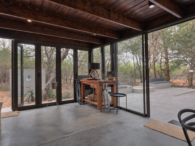 A modern covered patio featuring exposed timber beam ceilings, polished concrete floors, and expansive bi-fold glass doors opening to a natural bushveld setting.