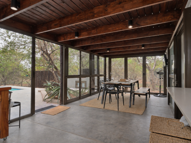 A spacious covered patio featuring exposed timber beams, concrete flooring, and large sliding glass doors that open to a garden and pool area.