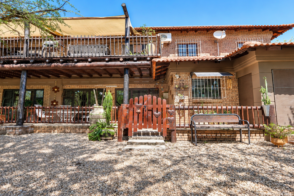 Rear view of a two-story brick house featuring a spacious timber balcony, shade sail, and a gravel-covered courtyard area with secure fencing.