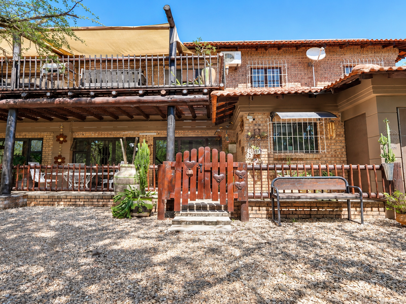 Rear view of a two-story brick house featuring a spacious timber balcony, shade sail, and a gravel-covered courtyard area with secure fencing.