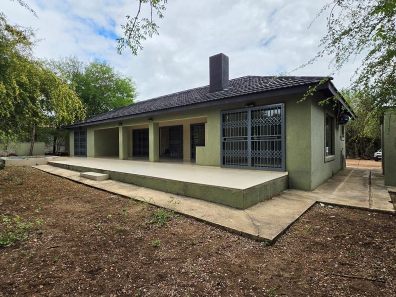 Rear view of the house featuring a spacious tiled patio, security gates, and a prominent chimney, set within a natural garden environment.