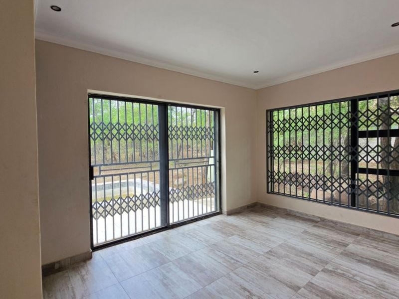 An empty living room featuring tiled flooring, recessed lighting, and large windows with security gates, offering a view of the outdoor area.