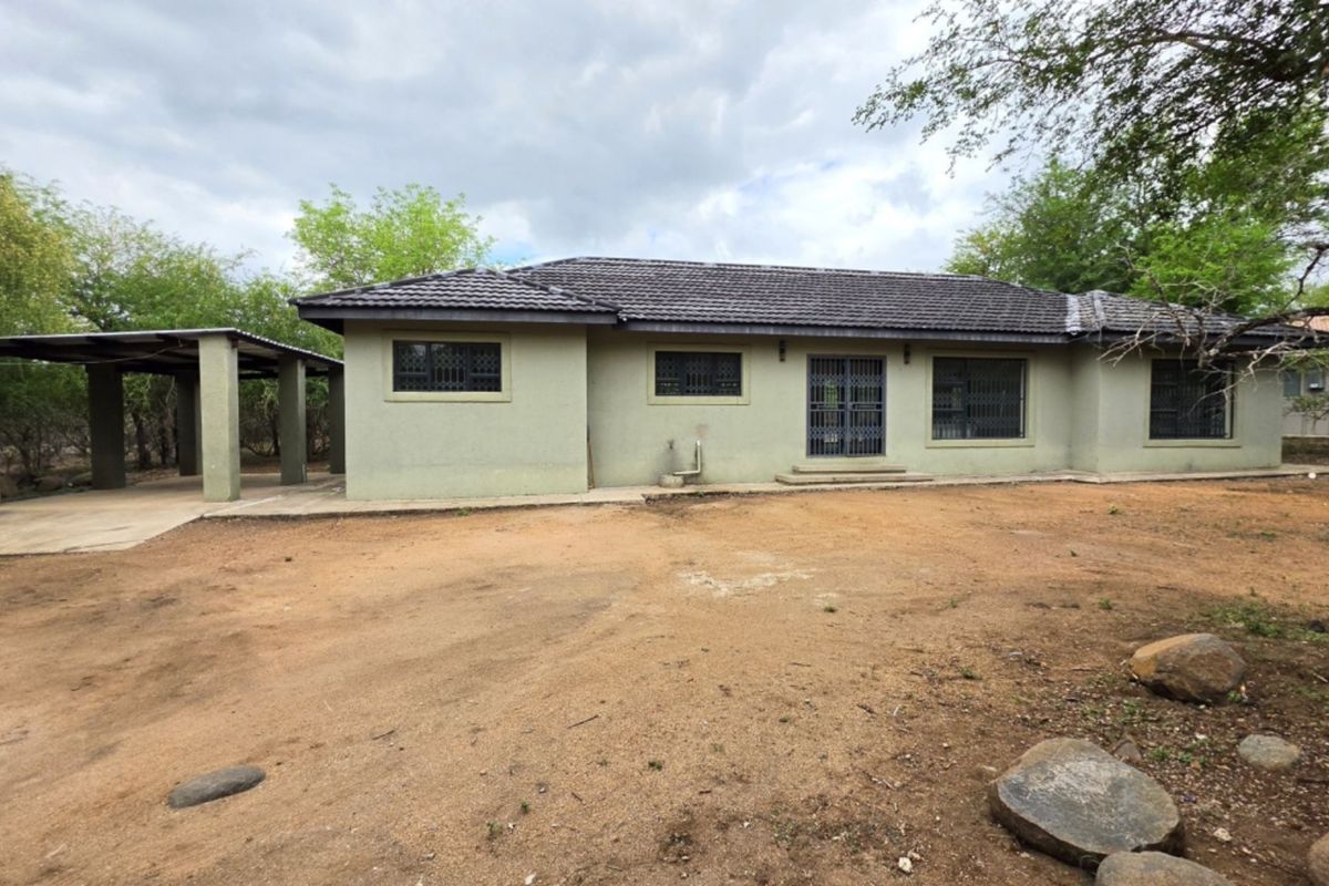 Rear view of a single-story house featuring a tiled roof, security gates, and an attached carport, set on a large unpaved plot.