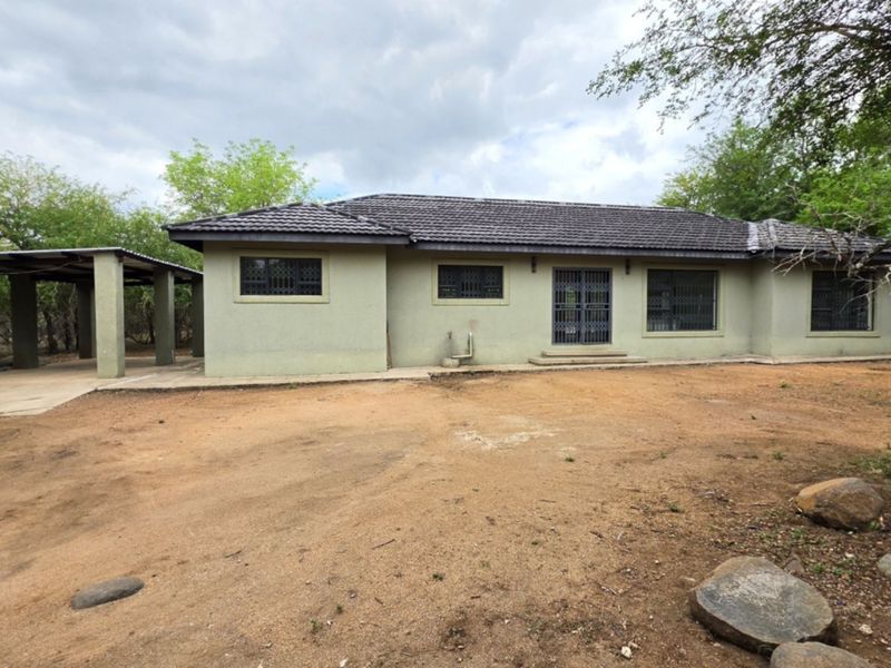 Rear view of a single-story house featuring a tiled roof, security gates, and an attached carport, set on a large unpaved plot.