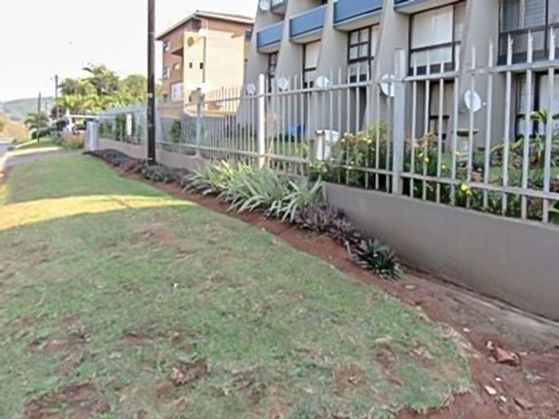 Exterior view of the flat complex featuring a metal security fence, concrete boundary wall, and maintained verge area.