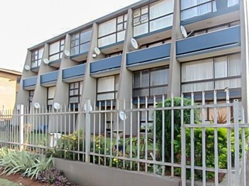 Exterior view of a multi-story apartment building featuring a concrete facade, balconies, and secure metal perimeter fencing.