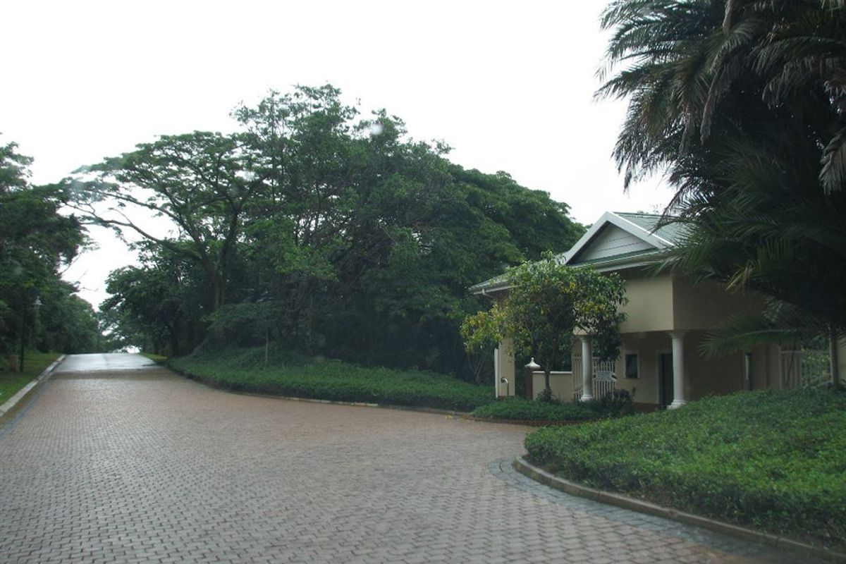 A view of the paved entrance driveway leading to a residential security gatehouse surrounded by mature trees and landscaped gardens.