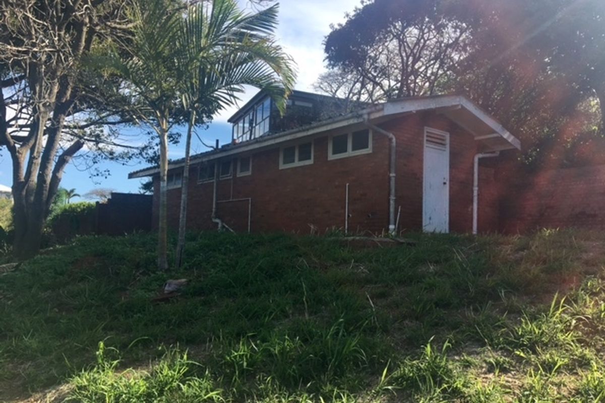 A view of the rear exterior of a brick residential structure featuring a pitched roof, guttering, and established surrounding trees on a sloped lot.