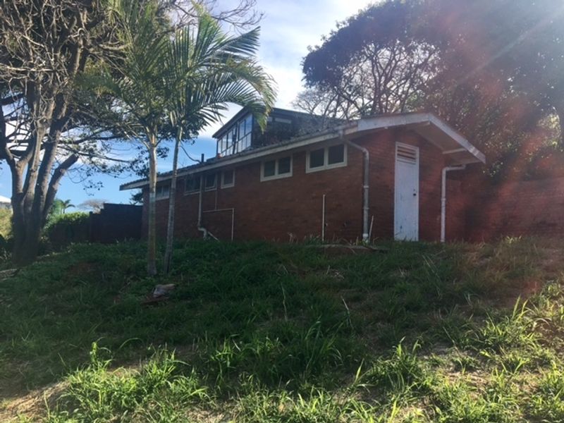 A view of the rear exterior of a brick residential structure featuring a pitched roof, guttering, and established surrounding trees on a sloped lot.