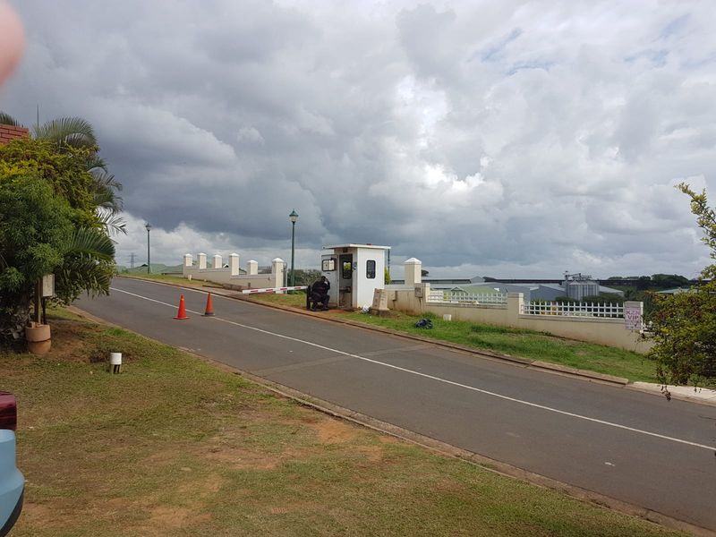 View of the estate entrance featuring a security guard booth, boom gate, and perimeter wall along the access road.