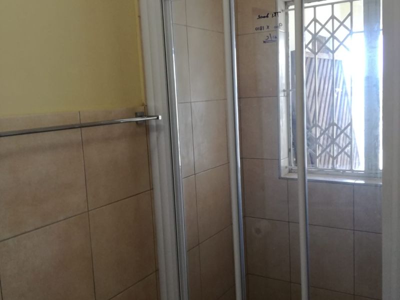 A functional bathroom featuring a glass shower enclosure, neutral wall tiling, and a window with security bars.