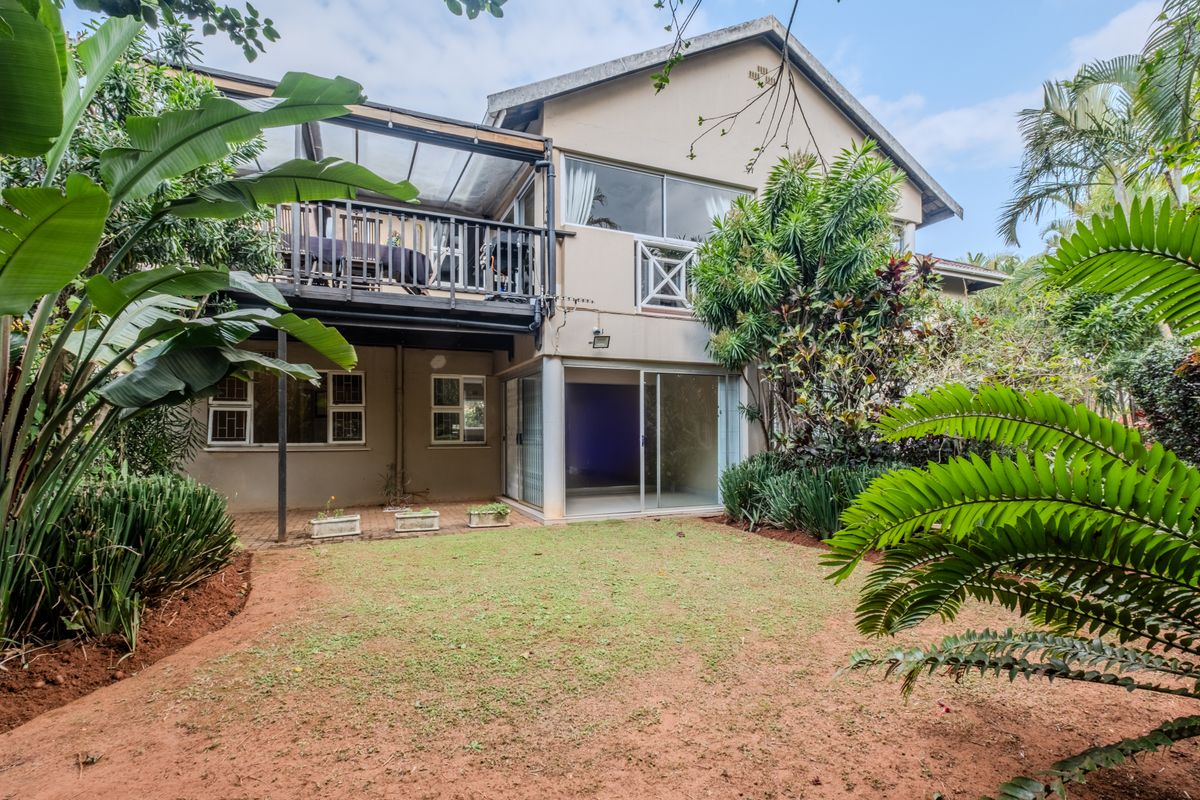 Rear view of the apartment building featuring a private garden, a paved patio area, and an elevated balcony with sliding door access.