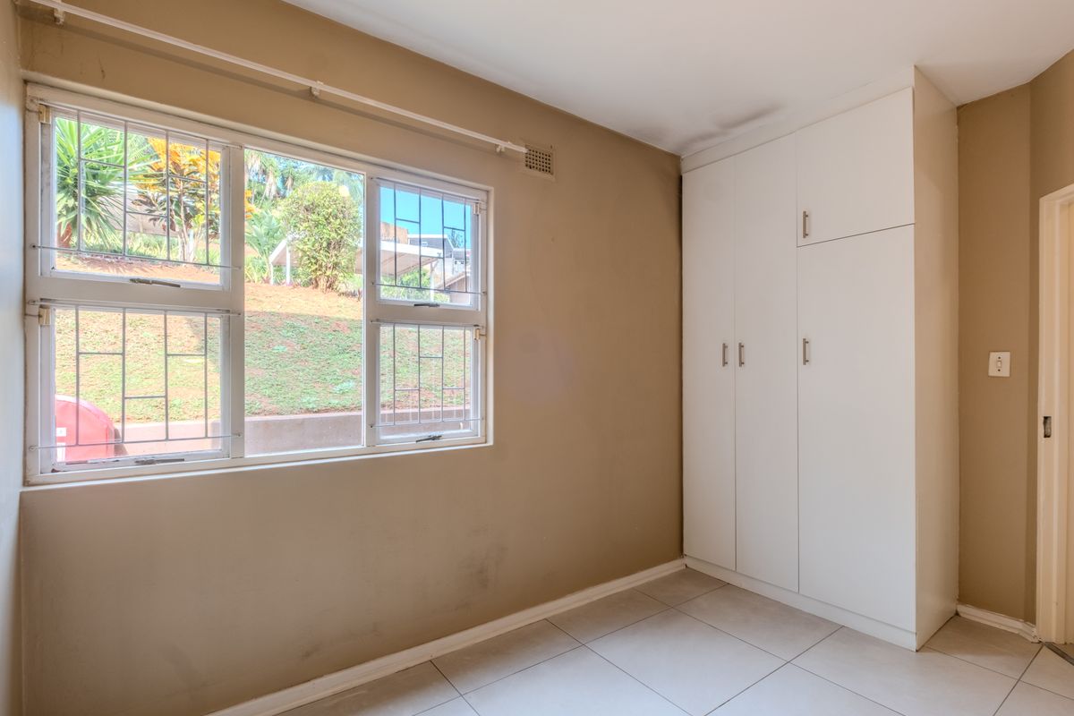 A bright, empty bedroom featuring tiled flooring, built-in cupboards, and large windows fitted with security bars.
