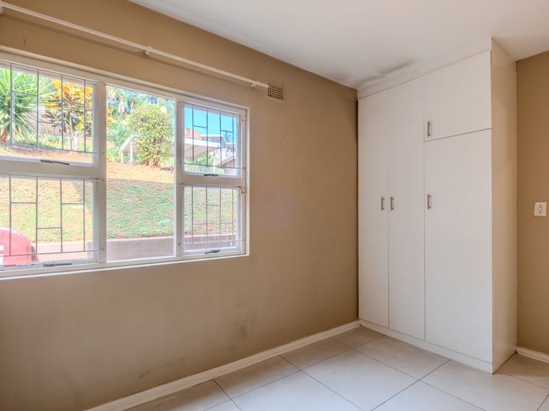 A bright, empty bedroom featuring tiled flooring, built-in cupboards, and large windows fitted with security bars.